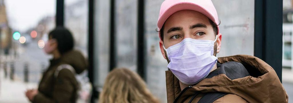 A teen wearing a pink hat and purple mask, waiting to catch a bus.