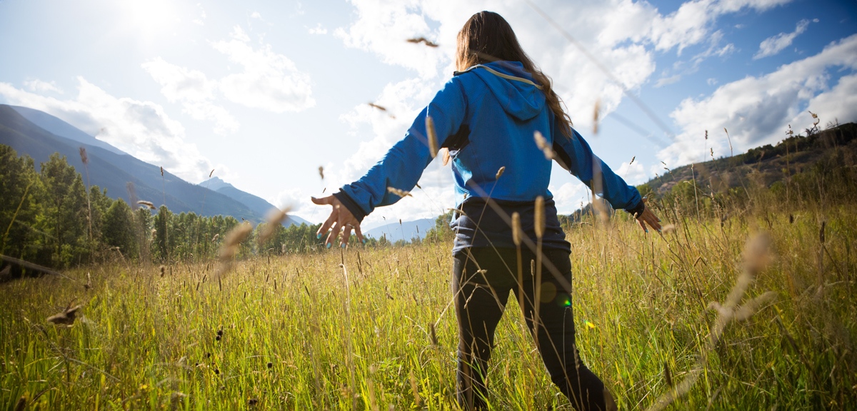 A photo of a youth walking through a field with their hands out catching the tall grasses.