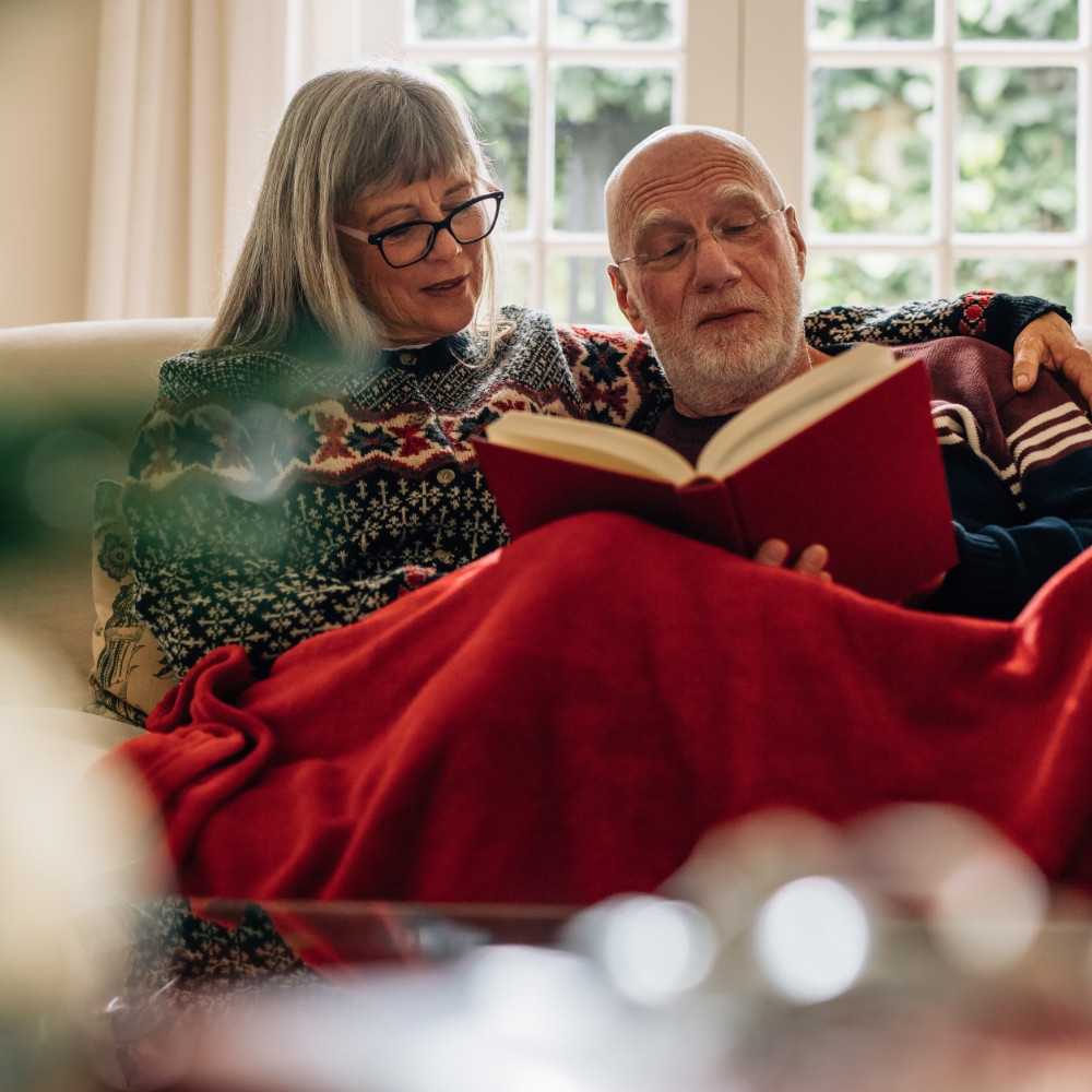 A photo of two people sitting on the sofa together reading a book.