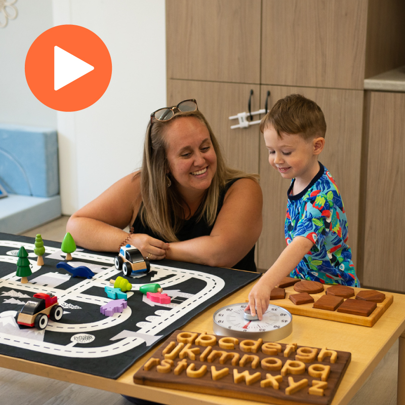 A caring adult and a young child are engaged in play-based learning at a table filled with inclusive and educational toys. The table features a toy road mat with colourful wooden vehicles and trees, a tactile wooden alphabet board, a shape puzzle, and a learning clock. The child is interacting with the clock while the adult offers encouragement and support. The setting is a bright, welcoming learning space designed to support early development. An orange play button icon appears in the top-left corner of the image.