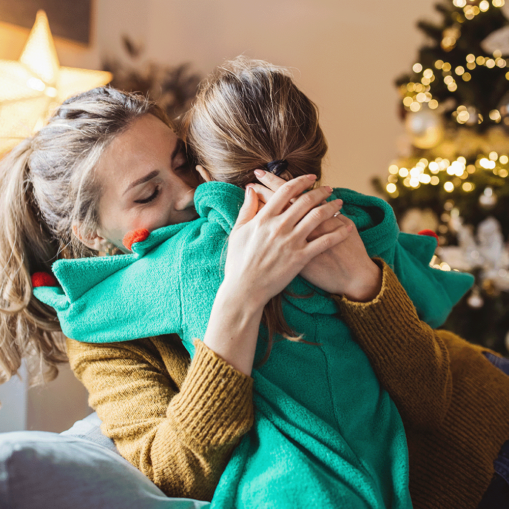 A mother and daughter hugging one another in their living room surrounded by holiday cheer.